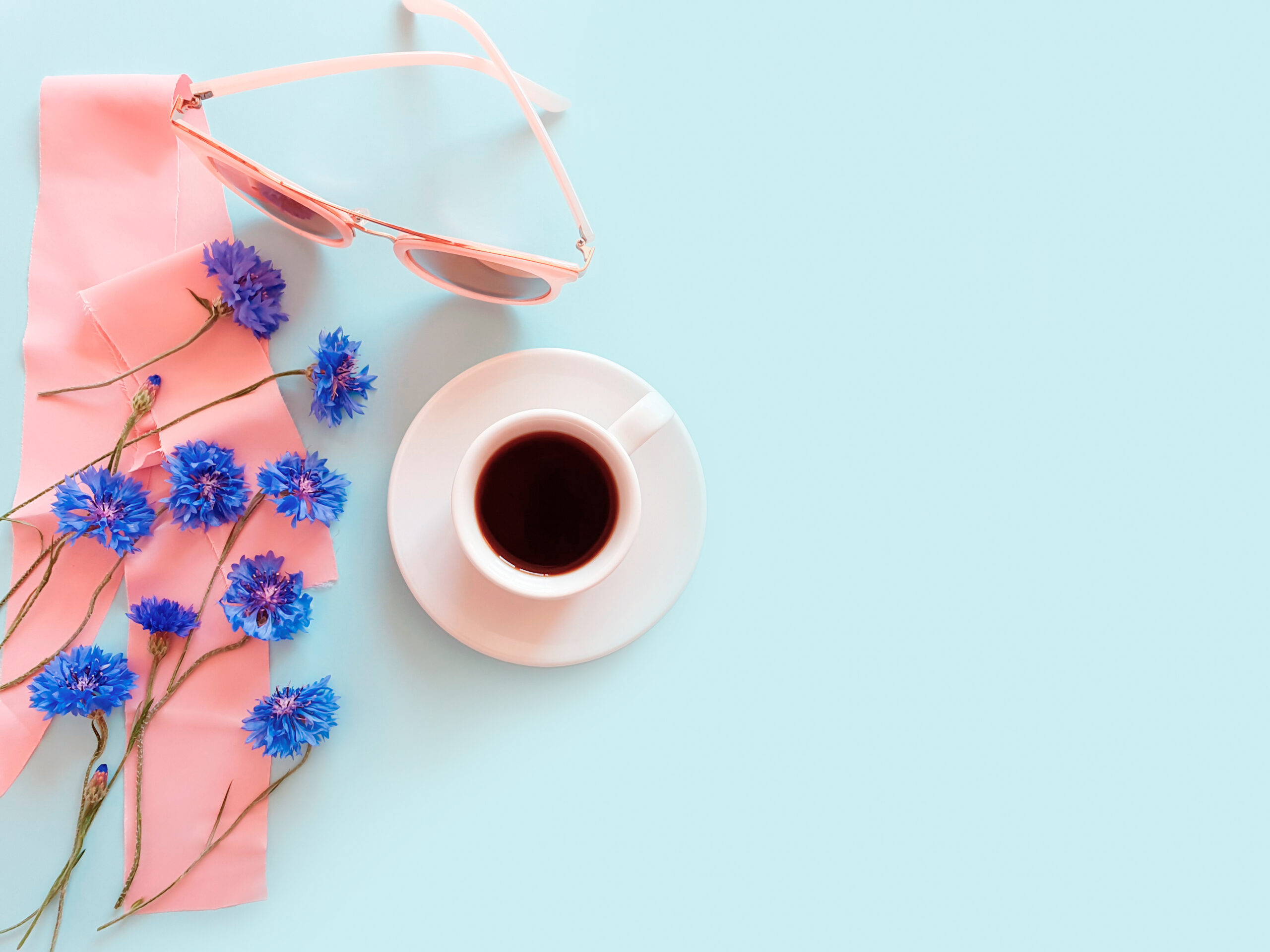 Top view of a cup of coffee with blue cornflowers, pink sunglasses and pink silk ribbon on a pastel blue background.